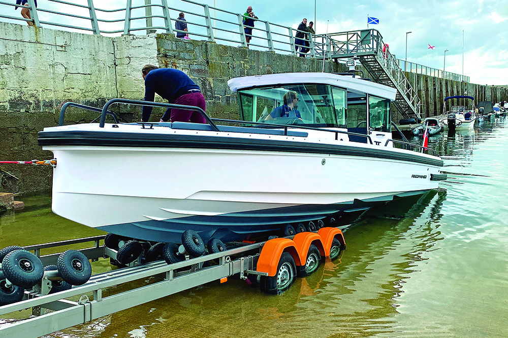 Axopar 28 Cabin launching from Vanclaes trailer at harbour slipway, showing gyro rollers and triple-axle support in shallow water.