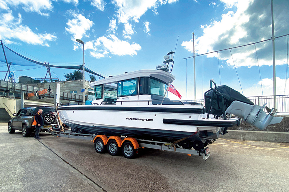 Axopar 28 Cabin on Vanclaes triple-axle trailer ready for towing, hitched to Land Rover Defender at marina slipway.