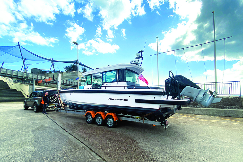 Axopar 28 Cabin on Vanclaes triple-axle trailer hitched to Land Rover Defender, parked at marina slipway ready for towing