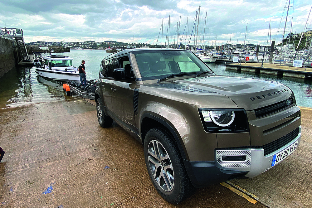 Land Rover Defender towing Axopar 28 on trailer at slipway, launching into marina with boats and pontoons in background.