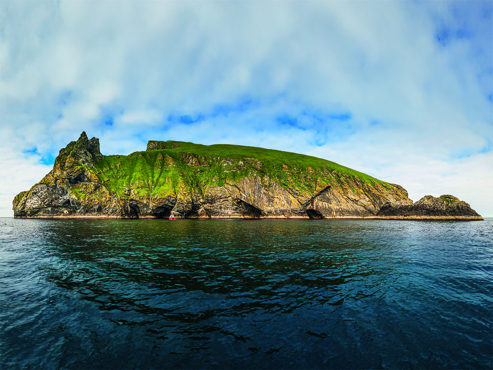 Green cliffs and sea caves of a St Kilda island rising sharply above calm Atlantic water under bright, broken cloud.