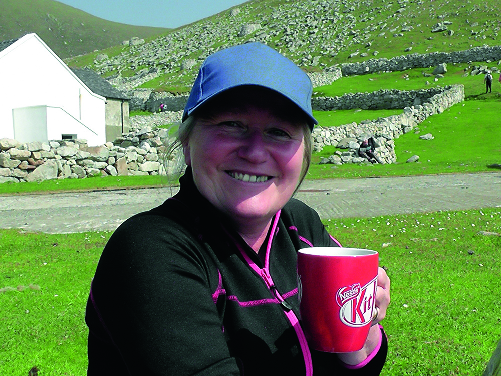 Jane smiling while holding a red mug, sitting among St Kilda’s stone walls and grassy slopes on a bright day.
