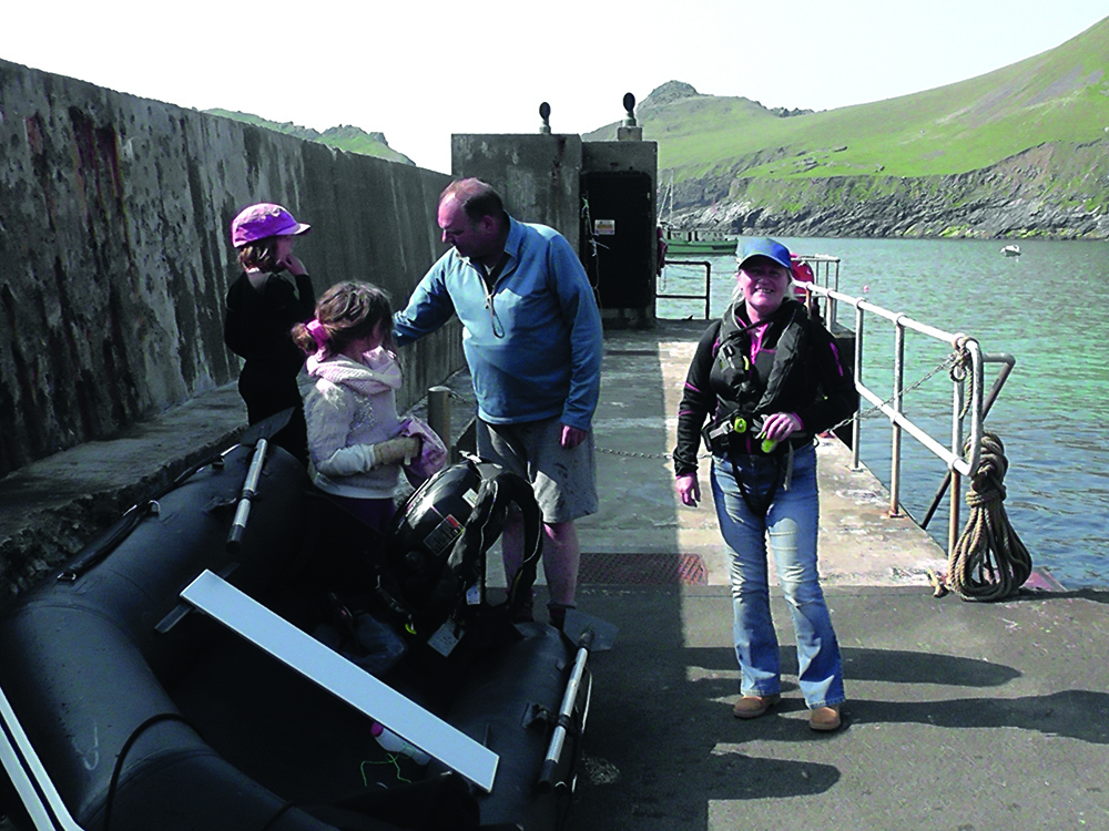 Family group standing on the pier at St Kilda beside a RIB, with green cliffs and clear water in the background.