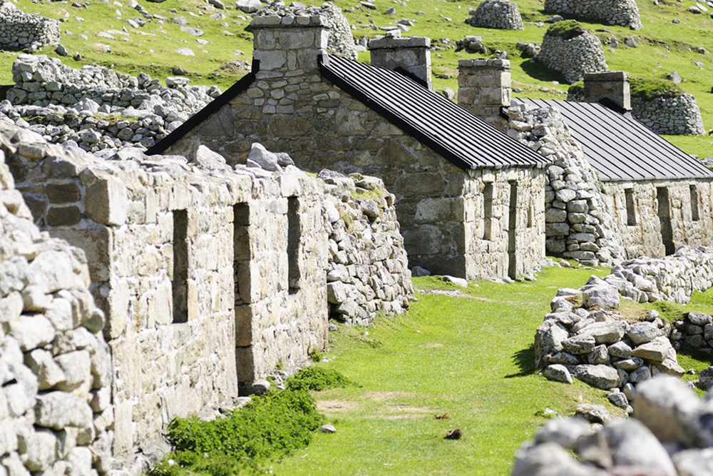 Ruins of cottages with green mown grass around and two restored cottages in the background on Hirta island