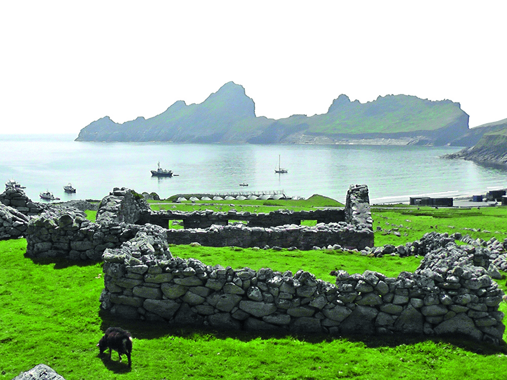 Stone ruins across the grassy slopes of Village Bay on St Kilda, with anchored boats and the dramatic outline of nearby sea stacks beyond.
