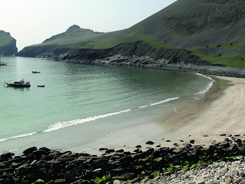 Curved sandy beach and calm turquoise water in Village Bay on St Kilda, with cliffs and anchored boats along the sheltered shoreline.
