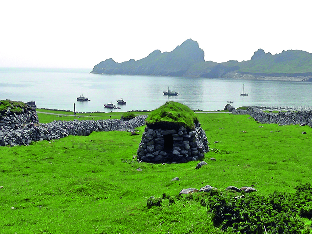Stone cleit with a grass roof overlooking Village Bay on St Kilda, with anchored boats and rugged sea stacks in the distance.