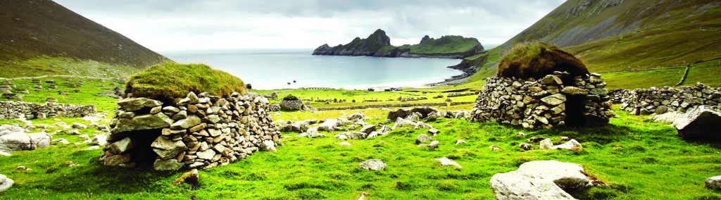 Stone cleits on Hirta overlooking Village Bay, with green slopes and jagged St Kilda sea stacks under a cloudy sky.