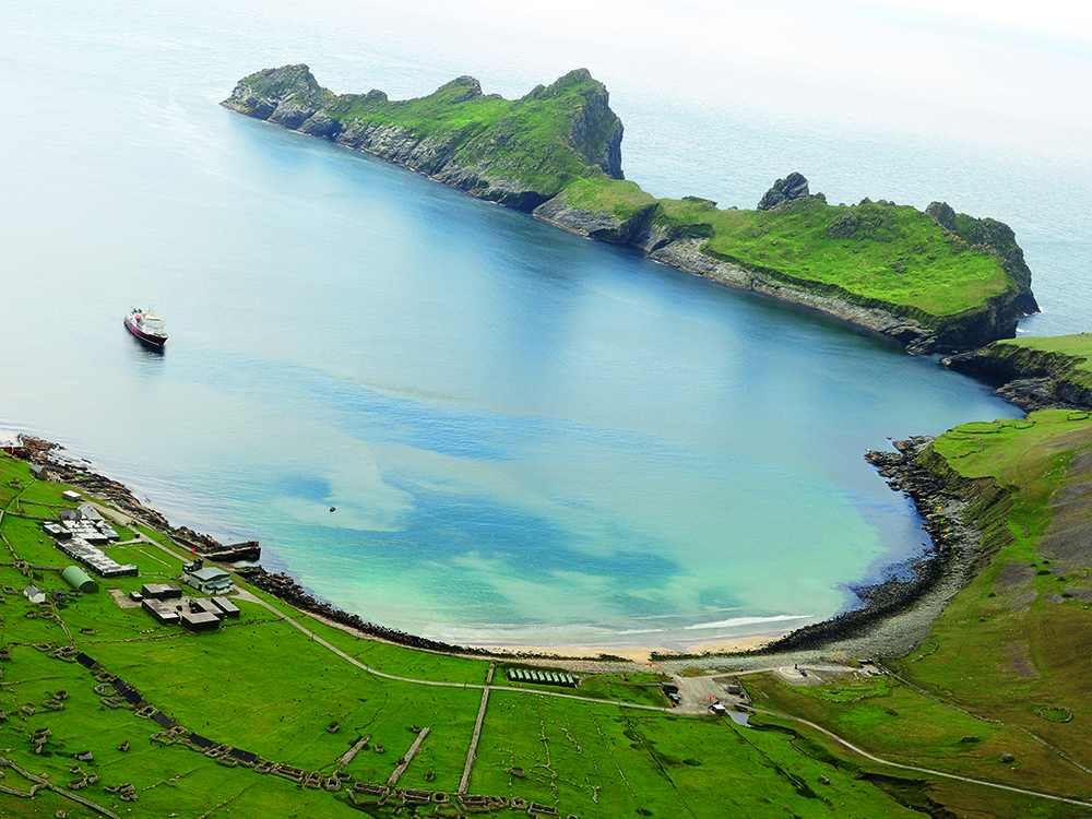 Aerial view of Village Bay on Hirta, showing turquoise water, green cliffs, and the scattered stone remains of the former St Kilda settlement.