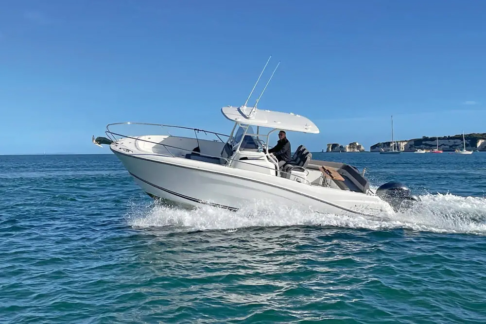 White centre console powerboat on plane, helmsman seated at the wheel, coastal cliffs and anchored yachts in background.