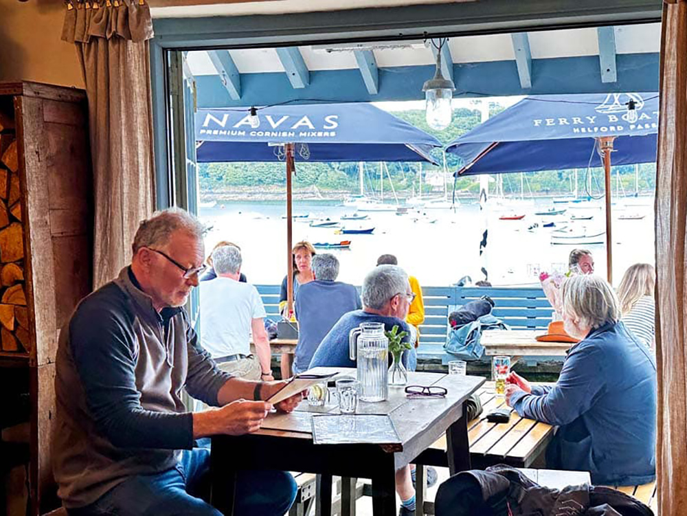 Customers enjoy food and drinks at the Ferry Boat Inn, overlooking the Helford River with moored boats visible through the open doorway.