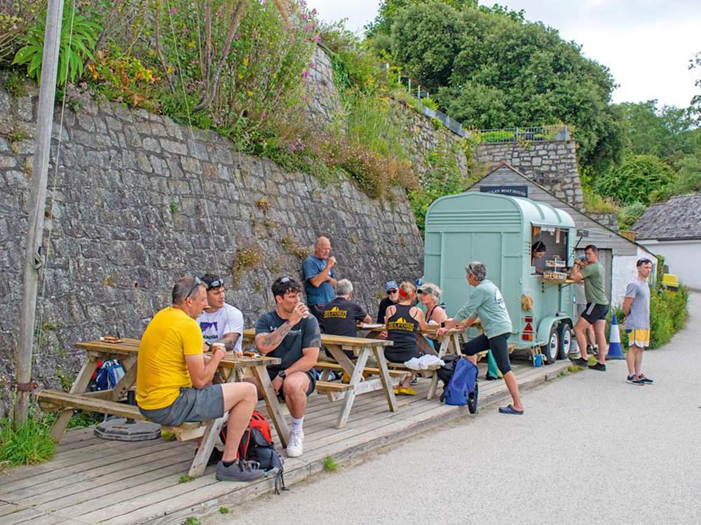 People enjoy coffee and snacks at outdoor picnic tables beside a converted horsebox café along the waterfront path at Helford Passage.