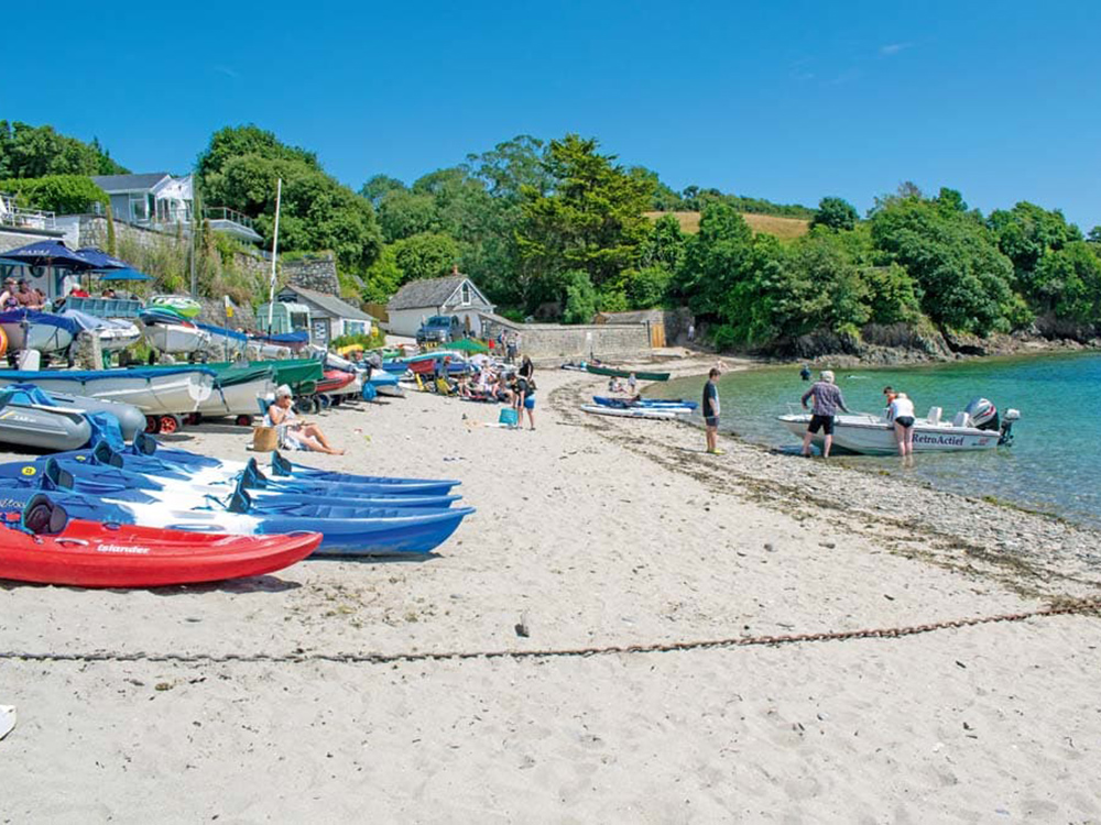 Visitors relax on the sandy beach at Helford Passage, with kayaks and small boats lined up beside clear turquoise water and wooded hills.