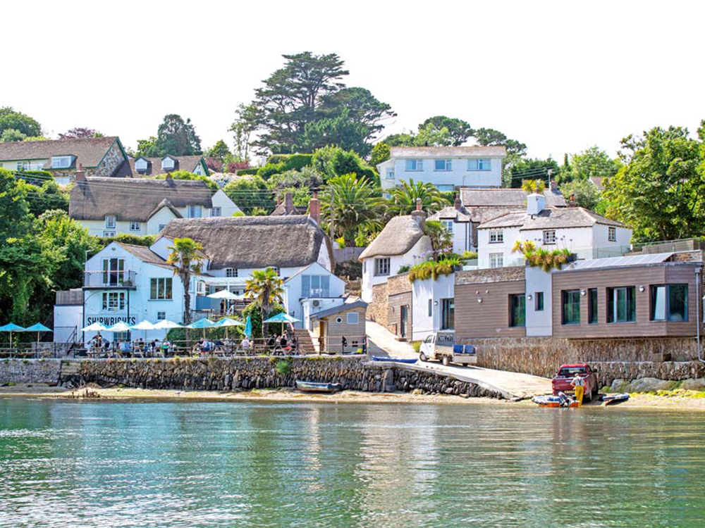 Waterside cottages and the Shipwrights Arms at Helford, with a fisherman unloading catch beside a slipway on a calm, sunny day.