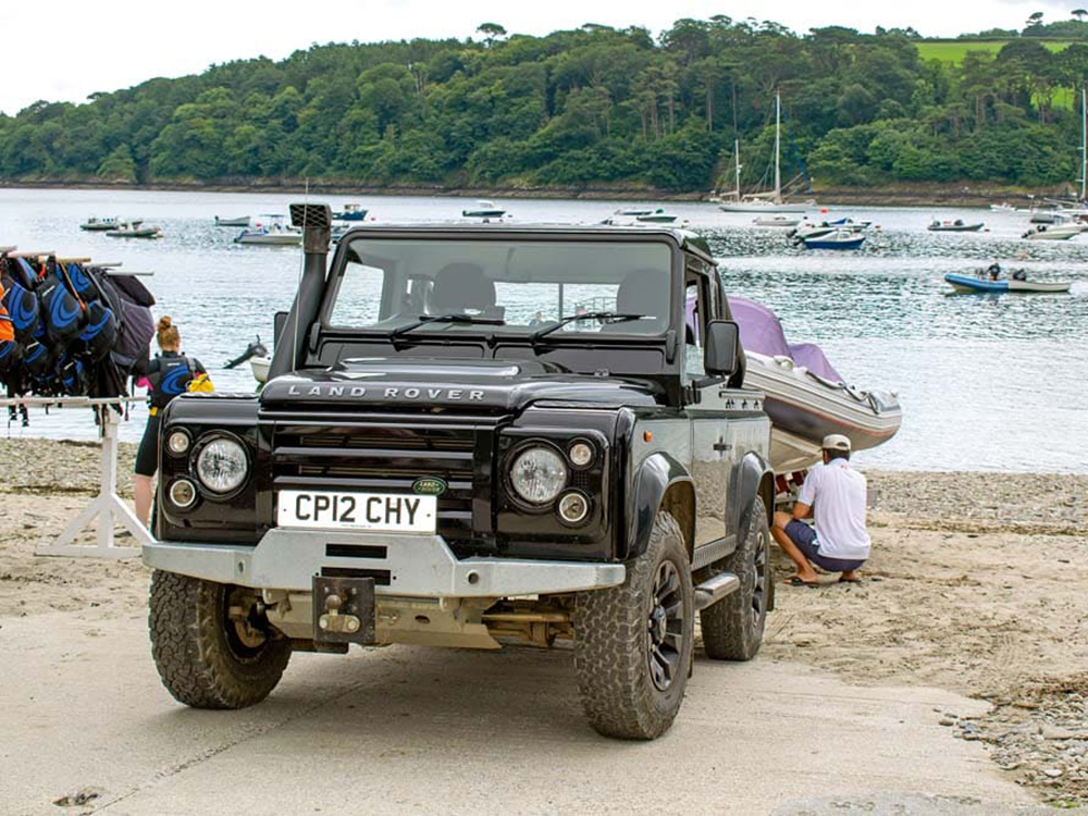 A Land Rover launches a RIB from the slipway at Helford Passage, with boats moored offshore and lifejackets hanging nearby.