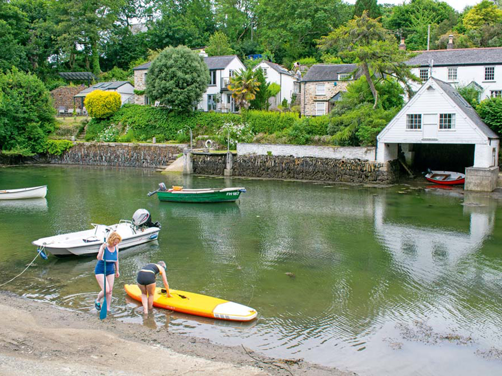 Two people prepare a paddleboard on a quiet Helford creek beside moored boats, stone cottages and a sheltered waterside boathouse.