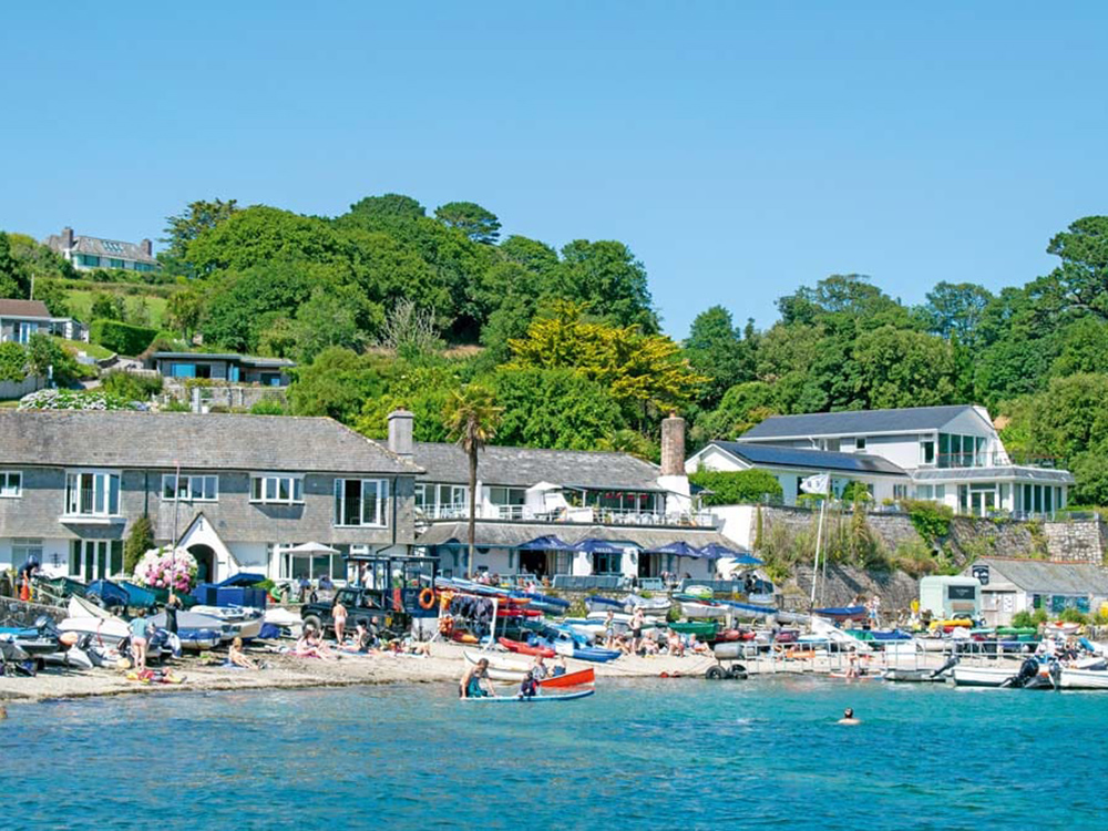 Helford Passage beach bustling with people, dinghies and small boats, backed by waterfront houses and lush green hills on a bright summer day.