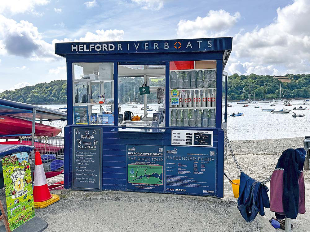 The Helford River Boats kiosk on the waterfront, offering ferry information, river tours and ice cream, with moored boats visible across the water.