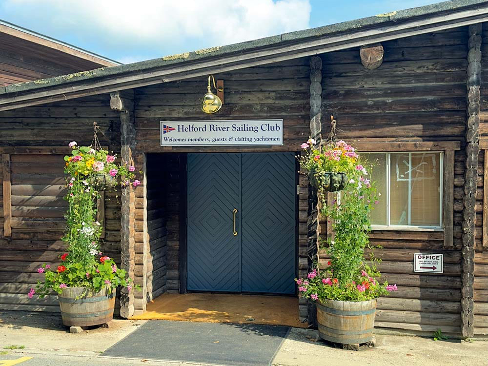 Entrance to Helford River Sailing Club, framed by wooden beams and colourful hanging flower baskets on a sunny day.