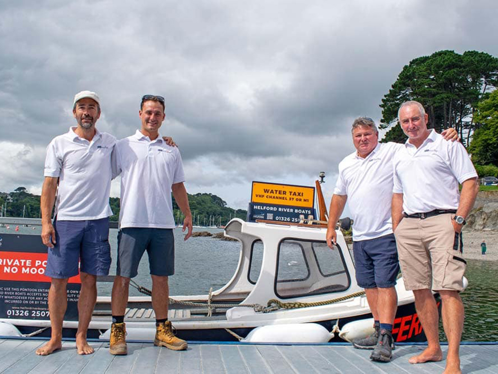 Four crew members stand on the pontoon beside the Helford River Boats water taxi, welcoming visitors against a backdrop of moorings and trees.