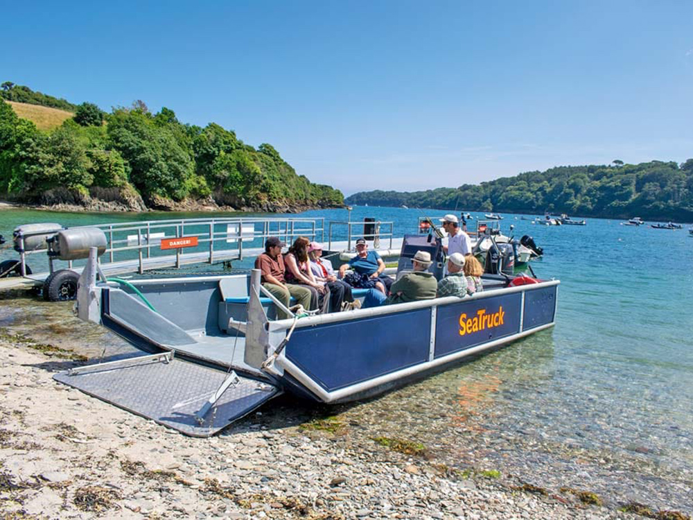 Passengers sit aboard the SeaTruck tour boat beached on a clear, shallow shore, with blue water and wooded hills across the Helford River.