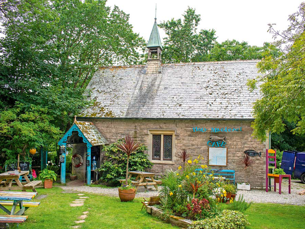 The Holy Mackerel Café set in a converted stone chapel, surrounded by colourful outdoor seating and lush garden plants.