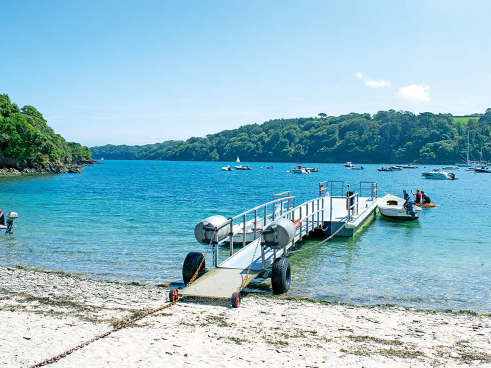 A floating pontoon extends from the beach at Helford Passage into clear blue water, with boats moored across the sheltered river.
