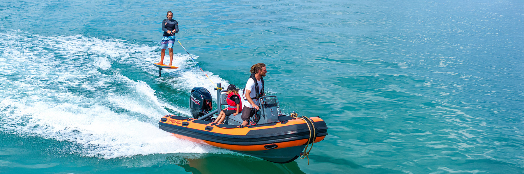 Orange Highfield RIB with man and child on board and another man wakeboarding off the stern