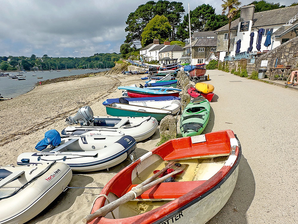 Small boats, dinghies and kayaks lined up along the waterfront path at Helford Passage, overlooking the sandy beach and moored vessels across the river.