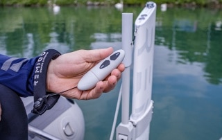 Hand holding RemigoOne waterproof remote control near electric outboard, with floating wrist strap and calm marina water in background.