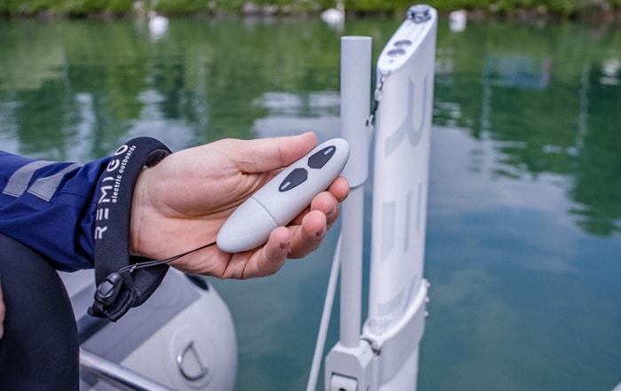 Hand holding RemigoOne waterproof remote control near electric outboard, with floating wrist strap and calm marina water in background.