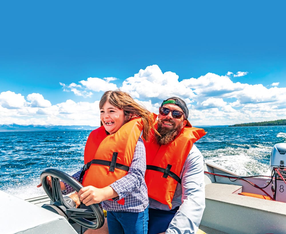 © Lisa5201 -iStockphoto Happy young girl wears a life jacket steering a small motor boat on a scenic lake with her family, togetherness in nature