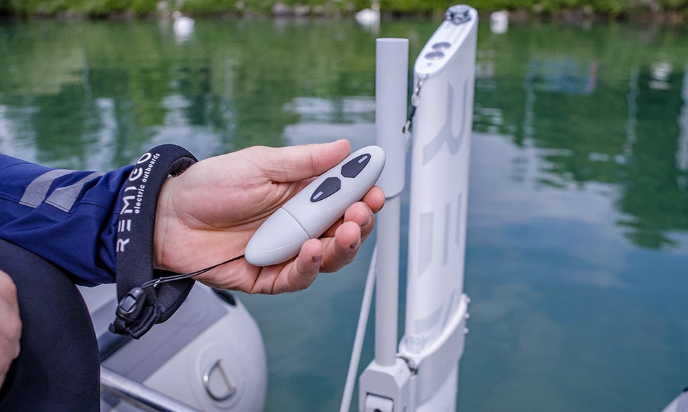 Hand holding RemigoOne waterproof remote control near electric outboard, with floating wrist strap and calm marina water in background.