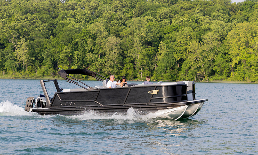 Alera Model M pontoon boat cruising on a lake with lush green forest in the background.