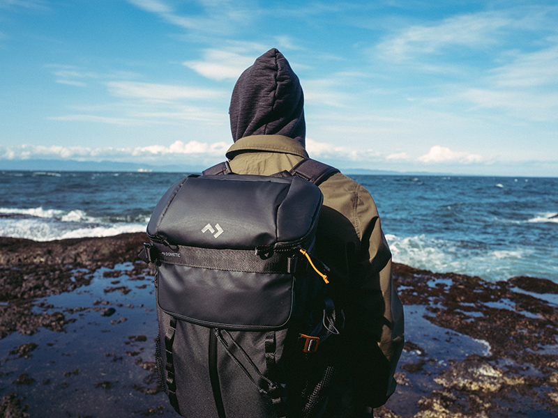 Person wearing a hooded jacket with a black Dometic Unrestricted backpack stands on a rocky shoreline overlooking the sea.