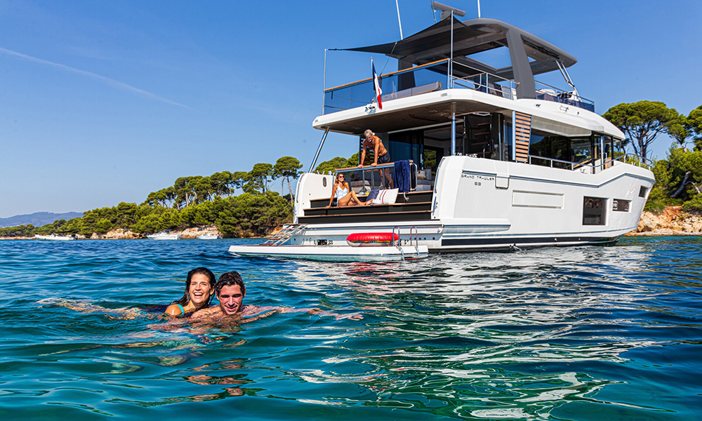 Couple swimming near anchored BENETEAU Grand Trawler 63 with family relaxing on the aft deck, set against a calm coastal backdrop.