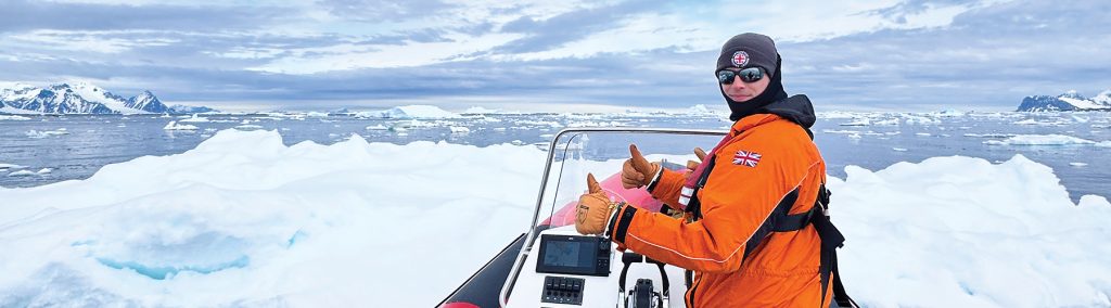 Ollie Tucker at the helm on a RIB looking back at the camera with icebergs in the background