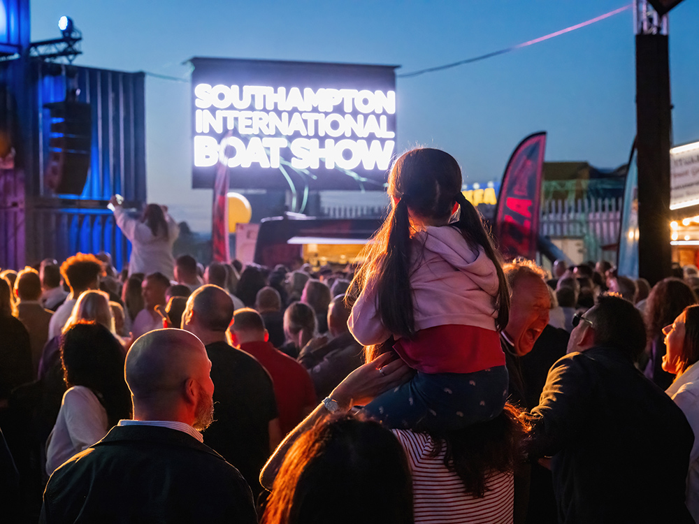 Crowds gathered at Southampton International Boat Show, with a child on shoulders watching evening entertainment.