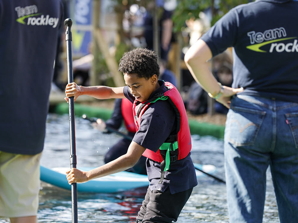 Young boy in a lifejacket learns paddleboarding under supervision at Southampton International Boat Show’s watersports activity zone.
