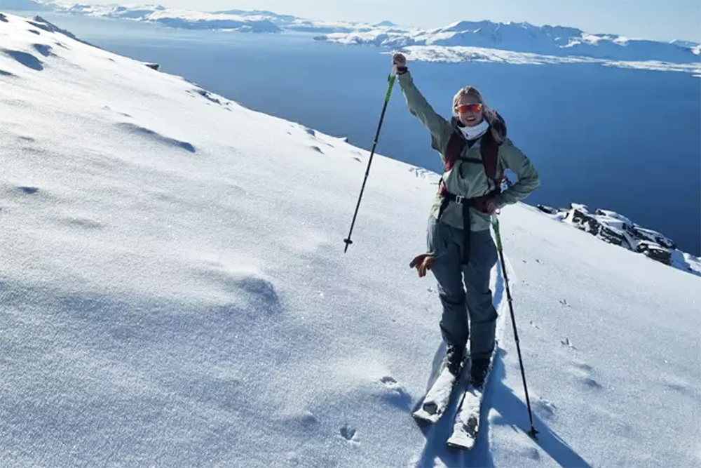 Female skier raising right arm in jubilation with snowy mountains behind.