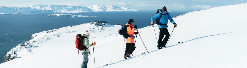 3 skiers climbing up snowy mountain with sea in background