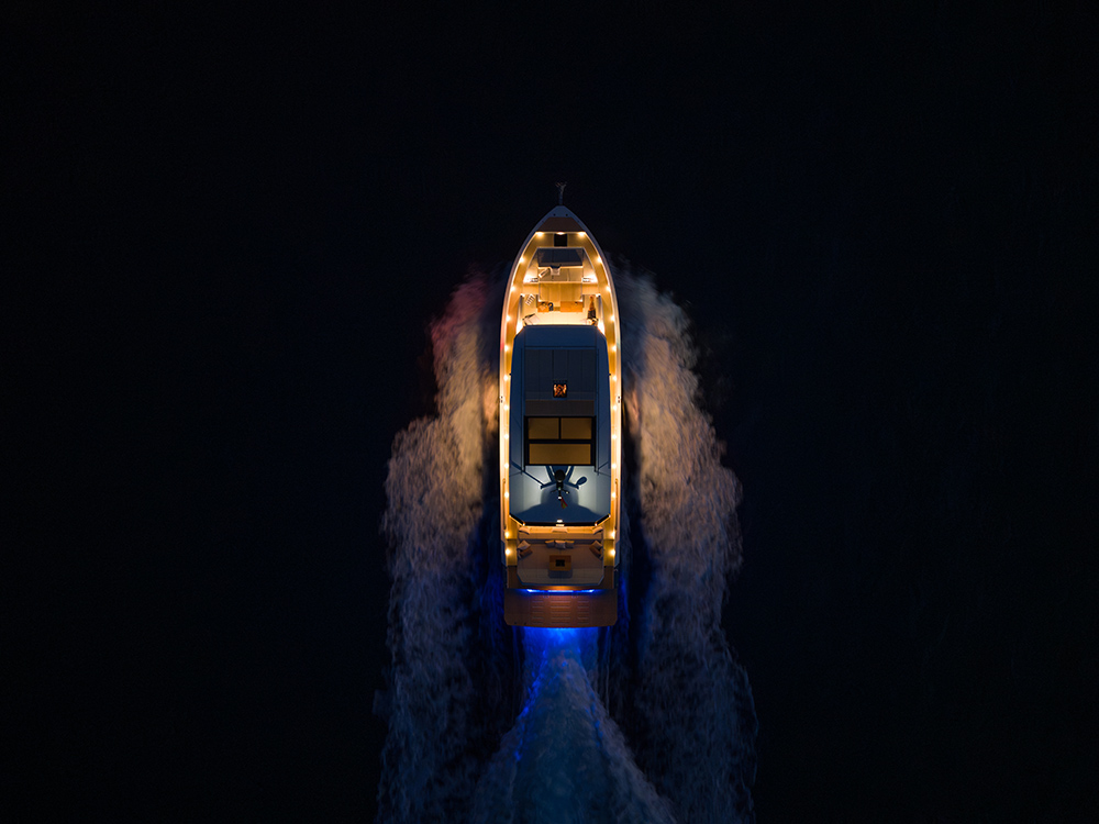 Overhead night view of De Antonio D60 yacht cruising, deck lights glowing and wake illuminated against dark water.