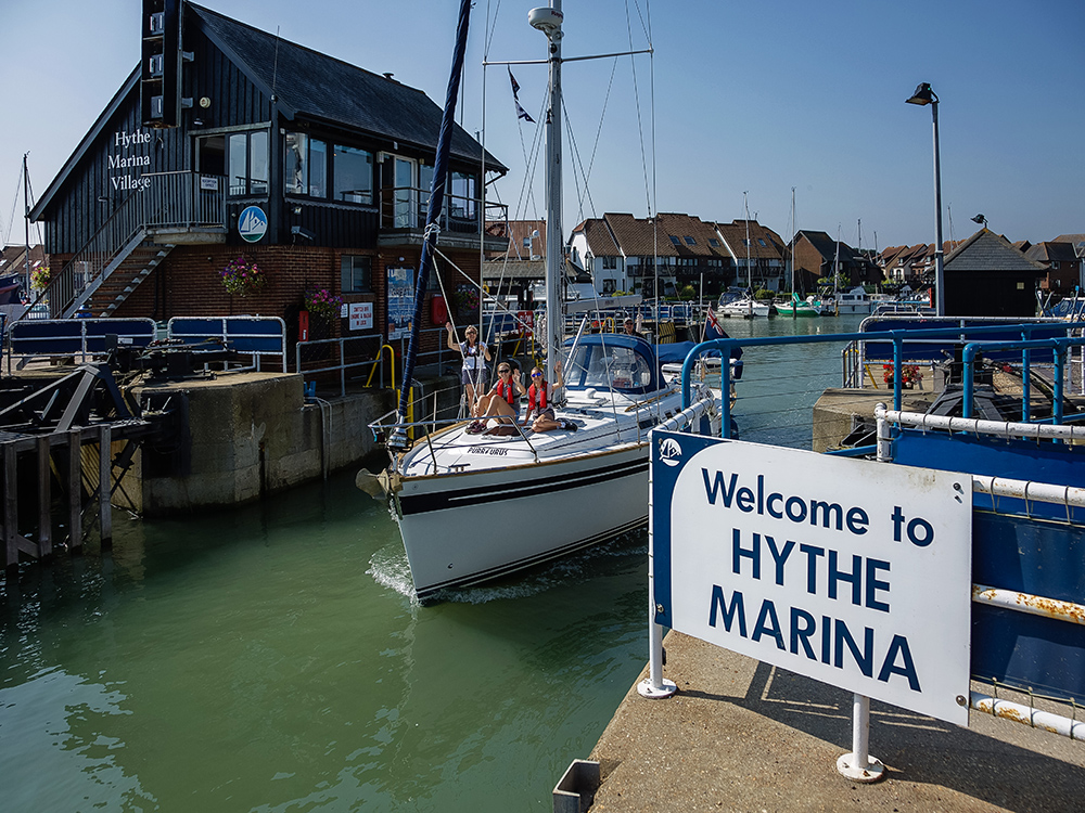 Sailing yacht entering Hythe Marina Village lock on a sunny day, with marina buildings, boats, and welcome sign in view.