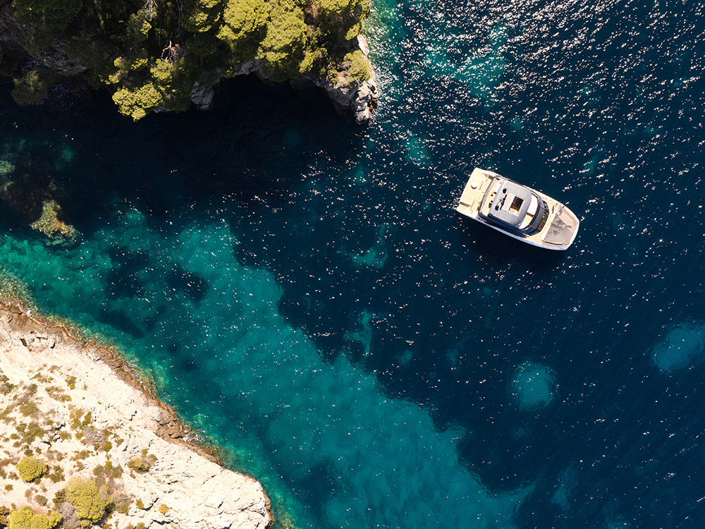 Aerial view of a Prestige M7 yacht anchored in clear turquoise water beside a rocky coastline.