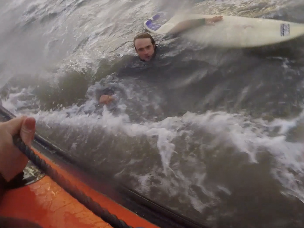 Surfer holding his board is pulled toward an orange RNLI lifeboat during a rough-sea rescue off Rest Bay, Porthcawl.