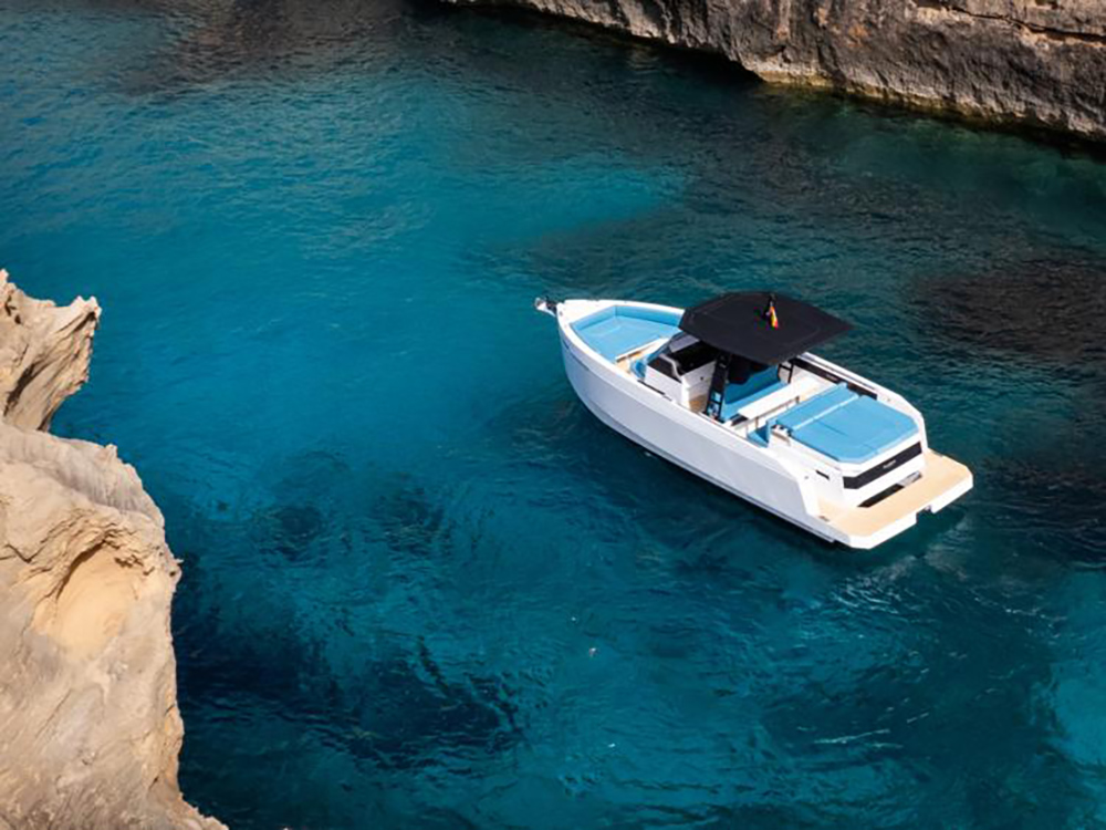 De Antonio yacht with Yamaha outboards anchored in clear turquoise water between rocky coastal cliffs.