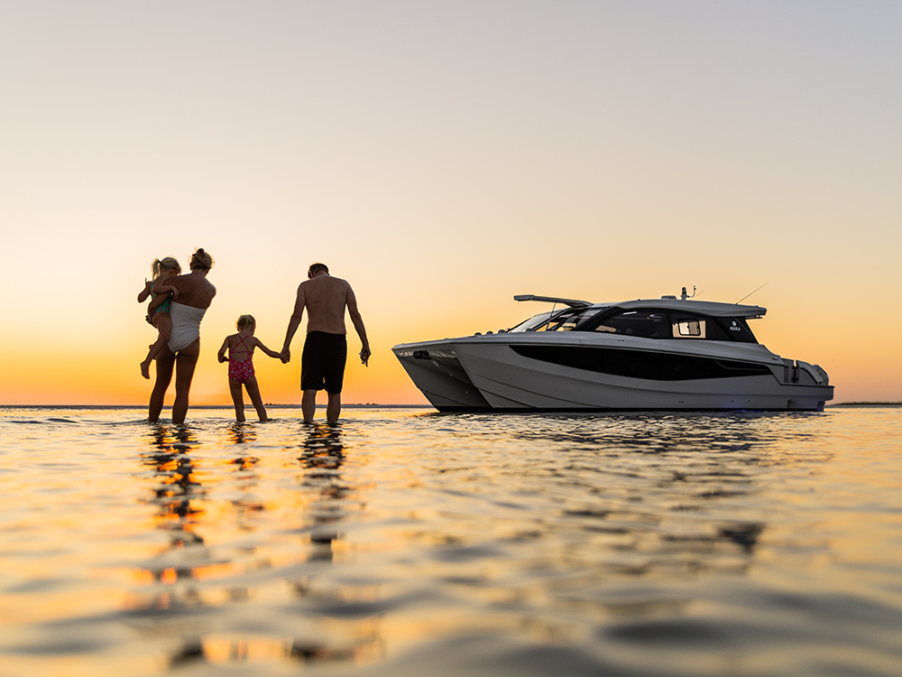 Family walking in shallow water at sunset with an Aquila 46 Coupe anchored nearby against a calm golden horizon.