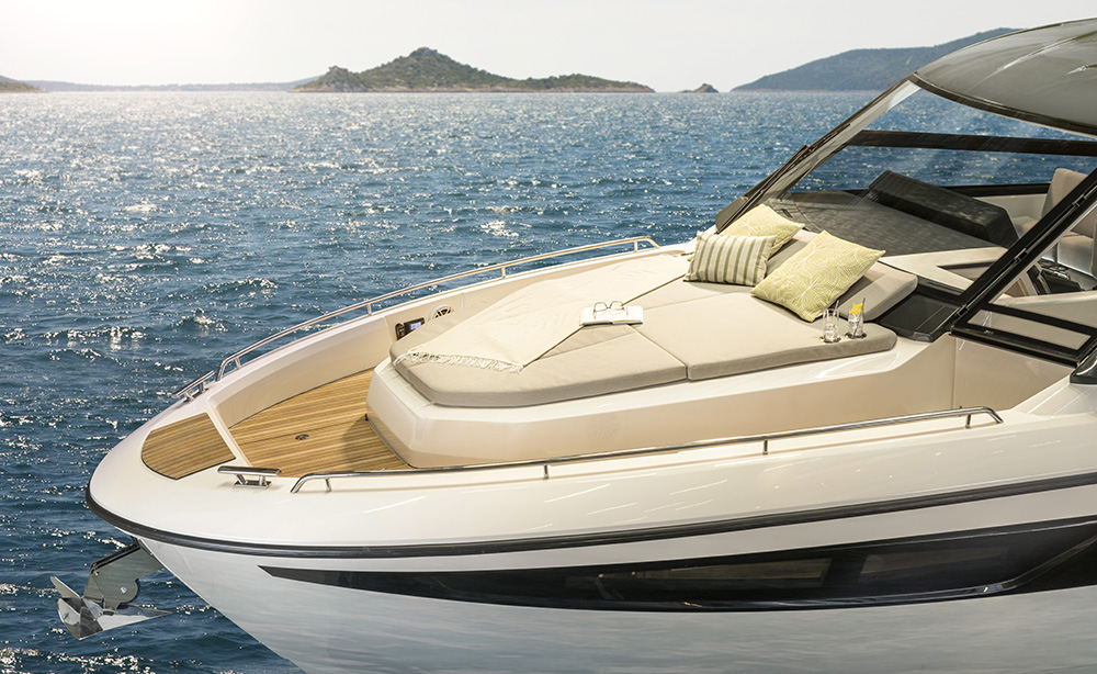 Bavaria VIDA 33 with teak foredeck and sunbed featuring cushions, blanket, and drinks, anchored on calm coastal waters.