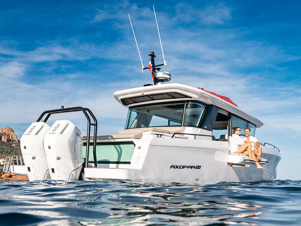 Axopar 45 with twin Mercury V10 400 hp engines afloat in calm water, two people relaxing on the aft deck under a clear blue sky.