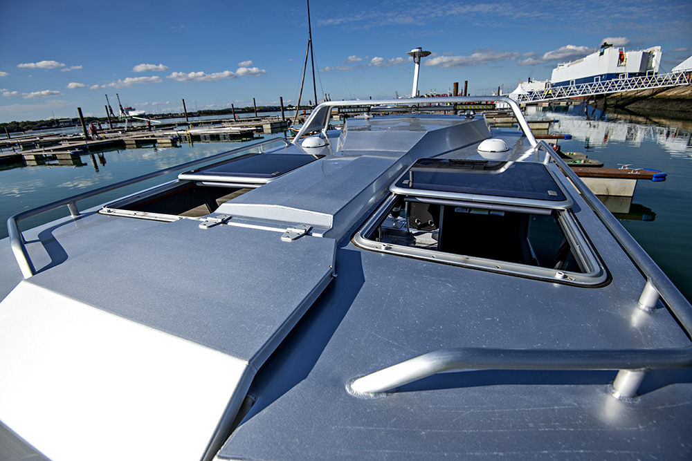 Ockelbo B25 CAB foredeck with open hatches and grab rails, reflecting sunlight beside marina pontoons under blue sky.